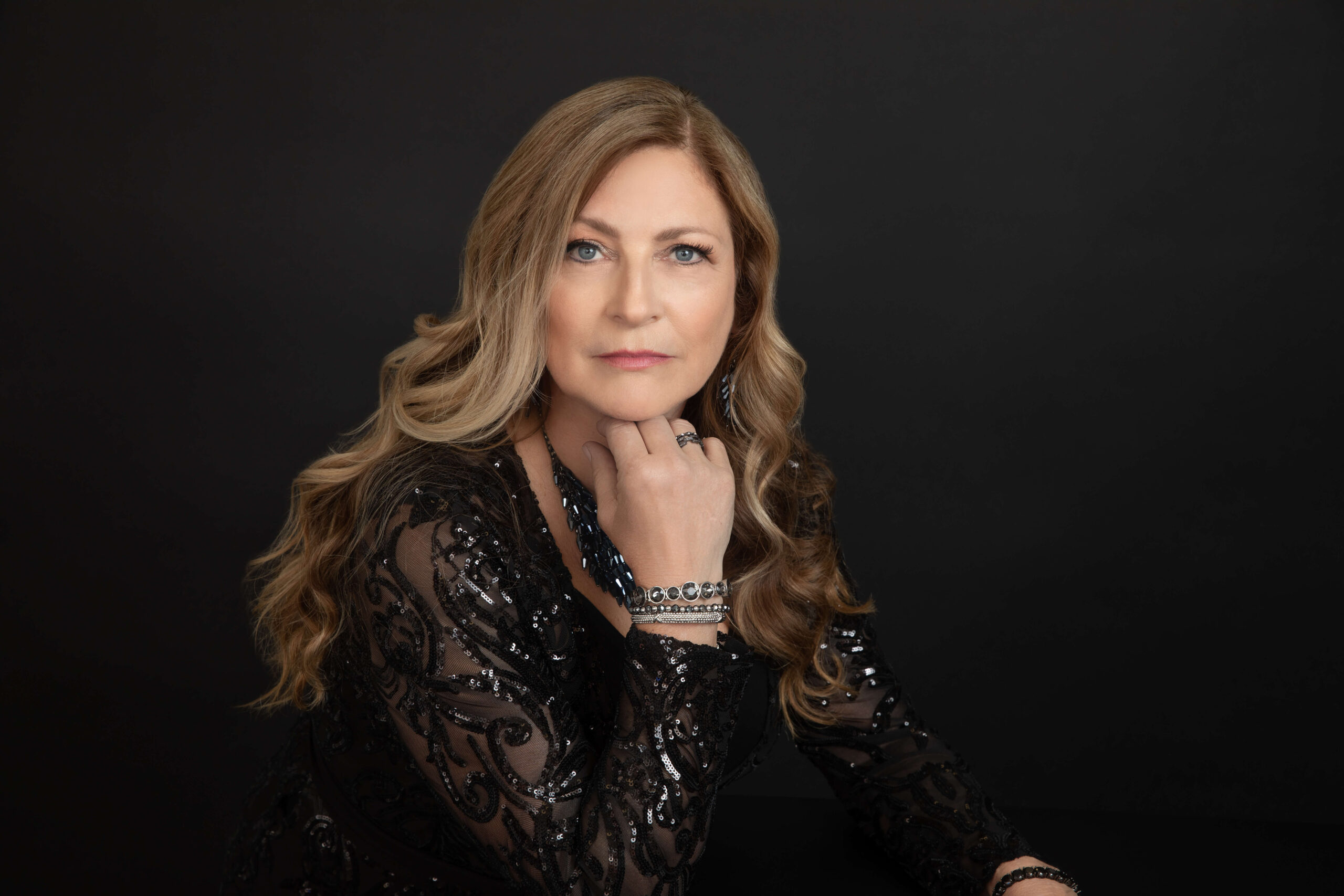 Woman with long, wavy hair wearing a black lace dress and jewelry, posing with her chin on her hand against a black background by Wenatchee Photographer Lynette