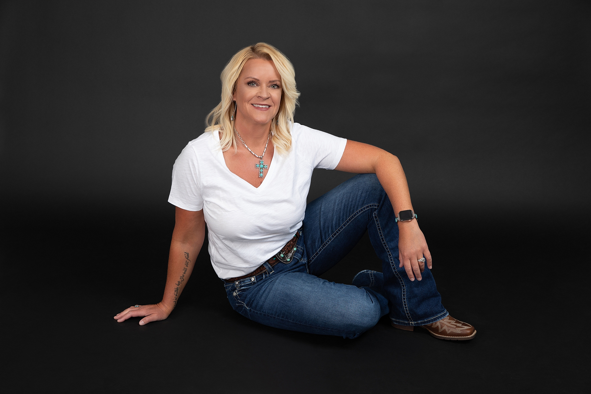 A woman with blonde hair sits on the floor against a black background, wearing a white t-shirt, blue jeans, and brown shoes. She is casually posed with one hand on the floor and the other resting on her knee, smiling at the camera in a Wenatchee Photography Studio.