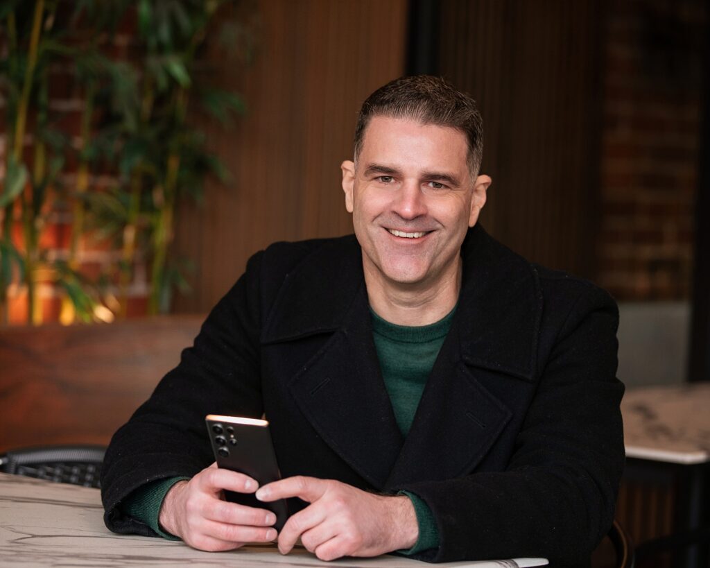 A person sitting at a table, smiling, holding a smartphone. They are wearing a dark coat and a green shirt. The background features a plant and blurred indoor elements. A branding or headshot photo.
