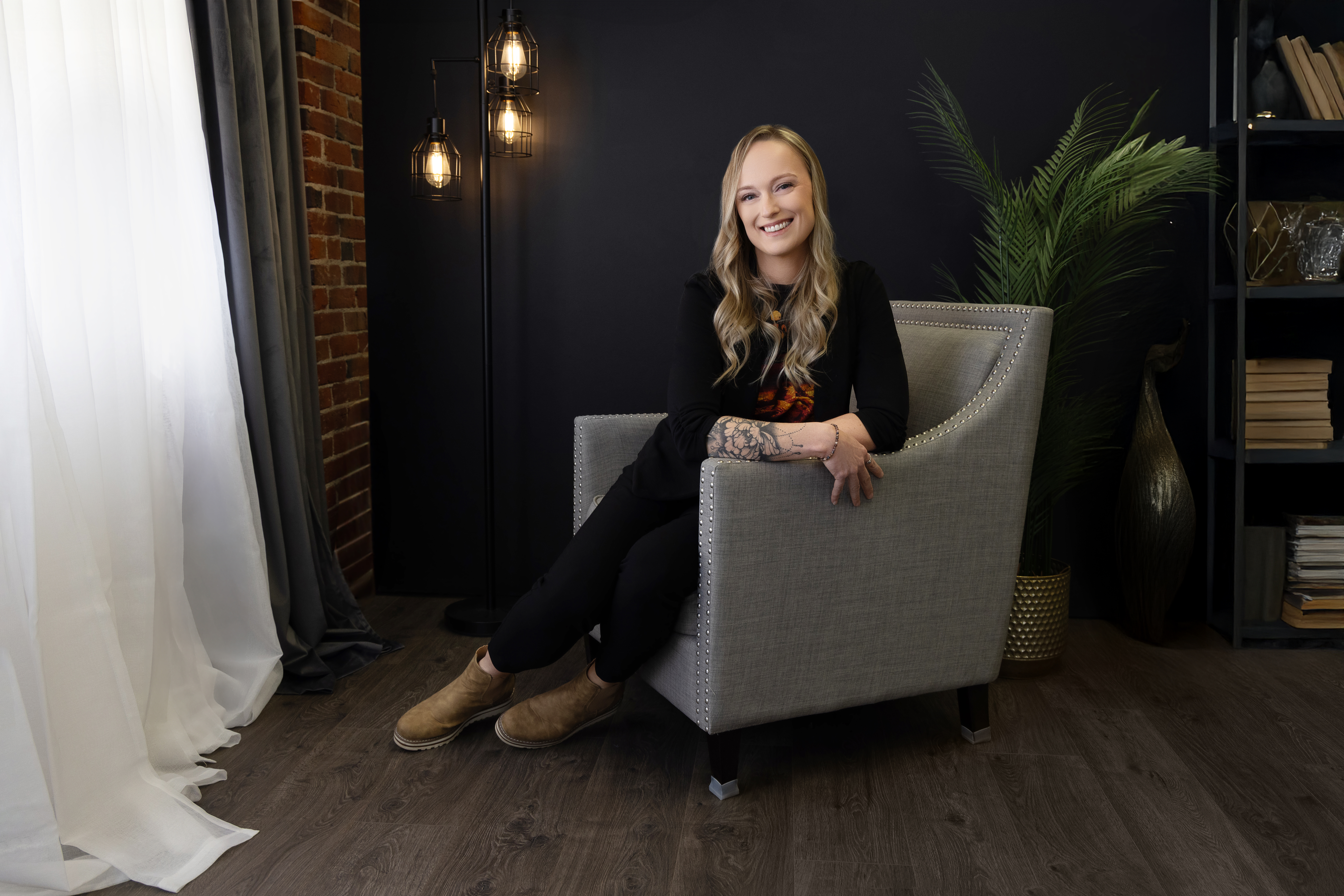 A smiling woman with long blonde hair wears a navy blazer, gray shirt, and ripped jeans. In one photo, she stands with hands at her sides; in the other, she sits on a gray chair, posing confidently in front of a dark background.
